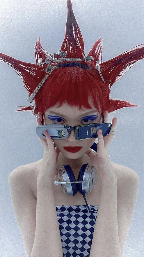 a woman with a red mohawk and checkerboard top is looking at the camera, she is wearing headphones and sunglasses.  she is looking at the camera.