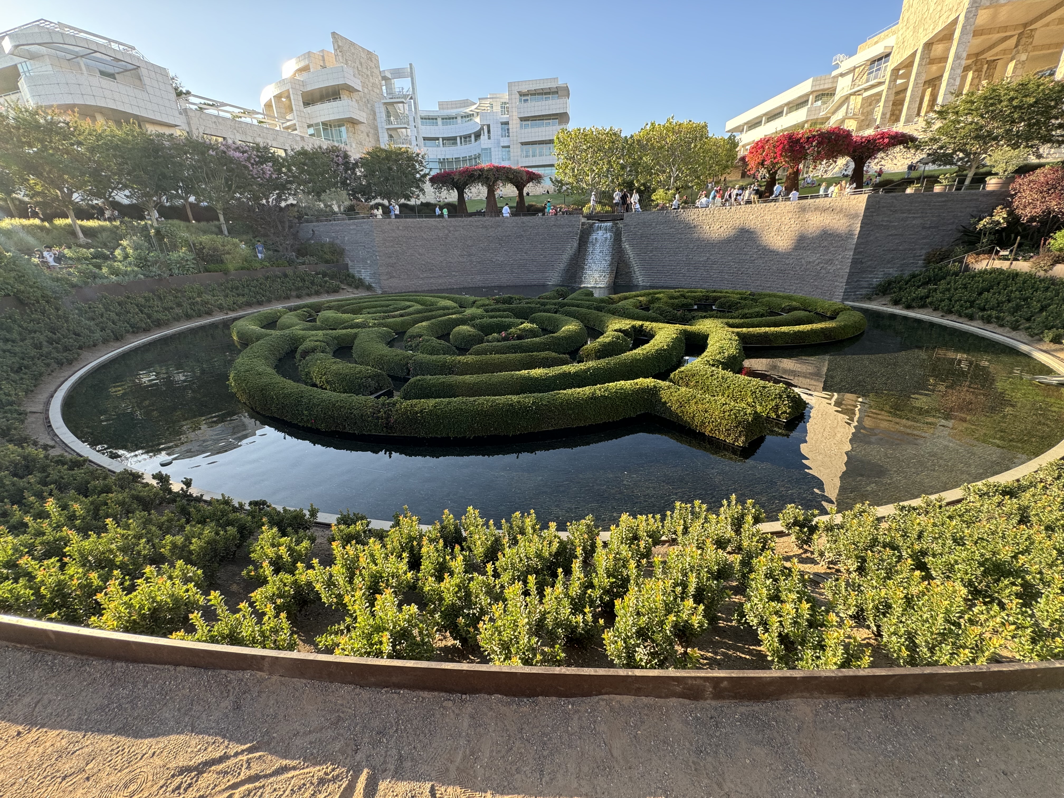a circular hedge maze sits in the middle of a fountain. the fountain is surrounded by a walkway and lush greenery. in the background, museum buildings and people can be seen enjoying the sunny day.