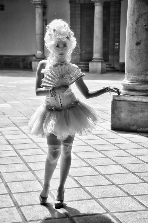 a black and white full shot of a young woman in a rococostyle costume, complete with a white wig, tutu dress, and handheld fan. she is posing outdoors on a stone patio with classical columns visible in the background. the lighting is natural, with some shadows indicating sunlight.  she is elegantly posed and her attire and style are evocative of a bygone era.