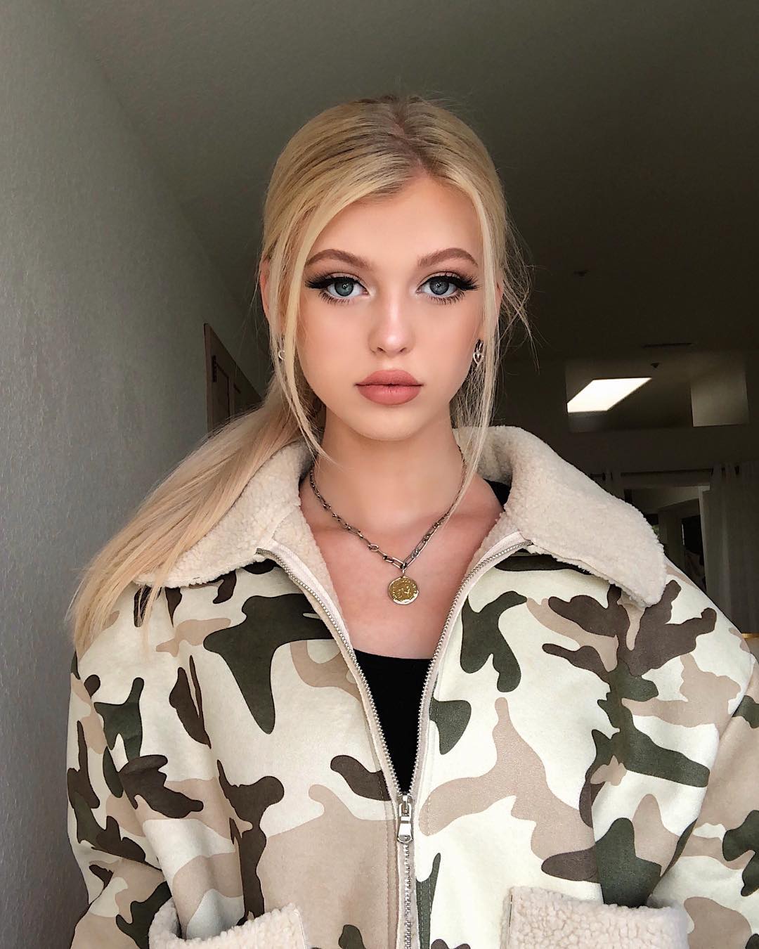 a portrait of a young woman with blonde hair and blue eyes. she is wearing makeup and a camouflage jacket with a fur collar. she has a necklace on and her hair is in a ponytail. she is standing indoors.