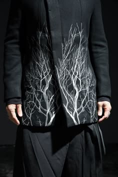 a man is standing in a studio with a dark background, wearing a black jacket with a white tree pattern. he is posing with his hands on his waist. he is stylish and modern, with a cool, elegant look.