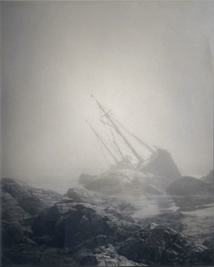 a monochrome image shows a shipwreck beached among large rocks, shrouded in fog. the remains of the ship's masts rise into the mistfilled sky. the scene is dominated by the heavy fog obscuring the background and the rough texture of the rocks in the foreground.