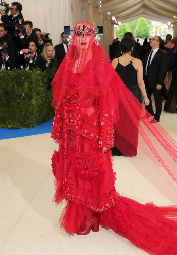 a woman in a bright red, elaborate dress and veil is standing on a white floor. the dress has layers of tulle and ruffles, along with detailed embellishments. she also has a sculptural headpiece with metal accents. the venue appears to be a formal event, possibly the met gala, indicated by the presence of other guests and photographers in the background. the red veil trails behind her, adding to the dramatic effect of the outfit.