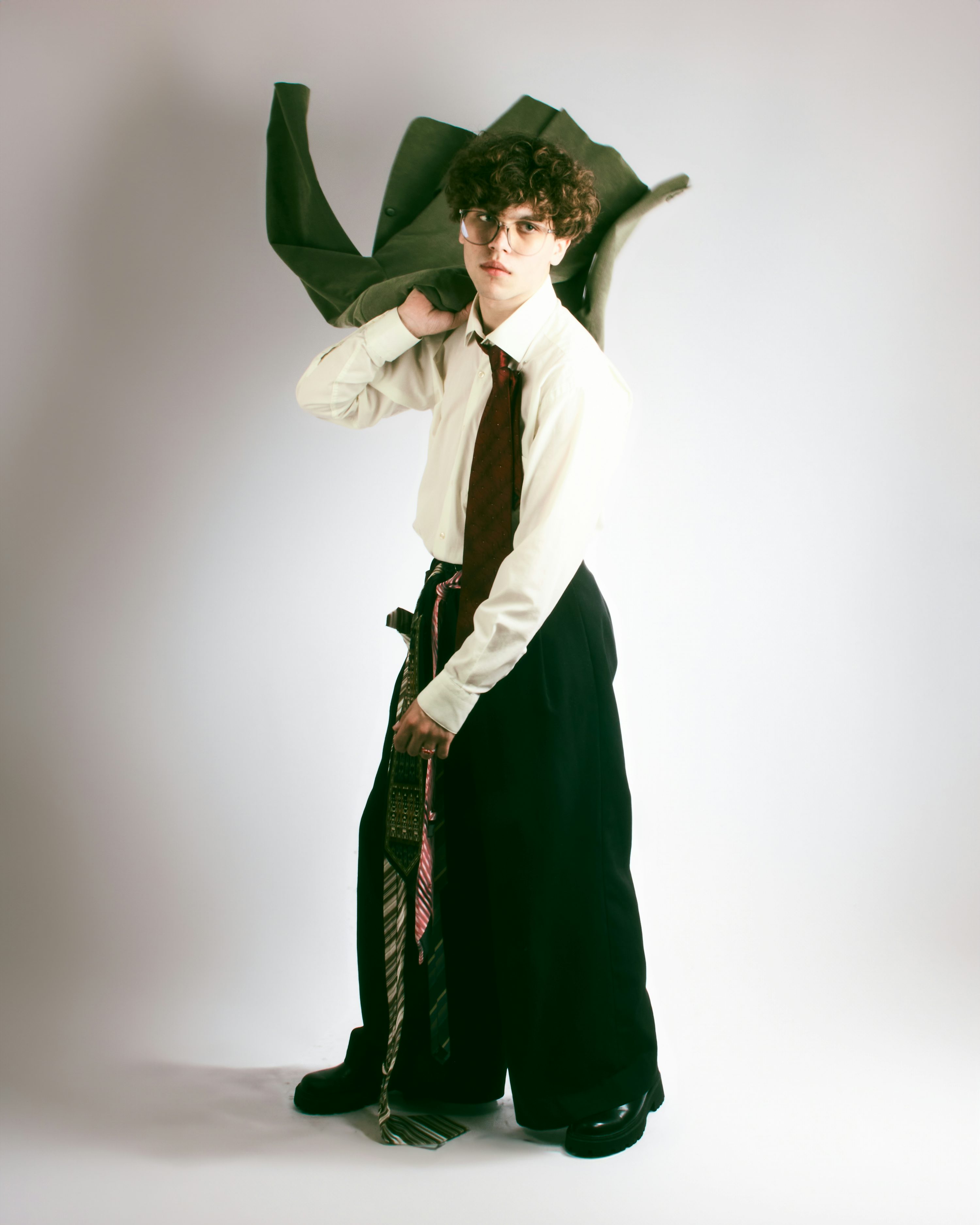the image is a stylish fashion portrait of a young man with curly brown hair wearing glasses. he is dressed in a white shirt, dark wideleg pants, and a dark tie.  he is carrying a green jacket over his shoulder,  and his overall appearance is fashionable and slightly quirky. the backdrop is a plain light grey, focusing attention solely on the model. the shot is fulllength, showing his entire outfit and pose. the model appears slightly serious in his expression.