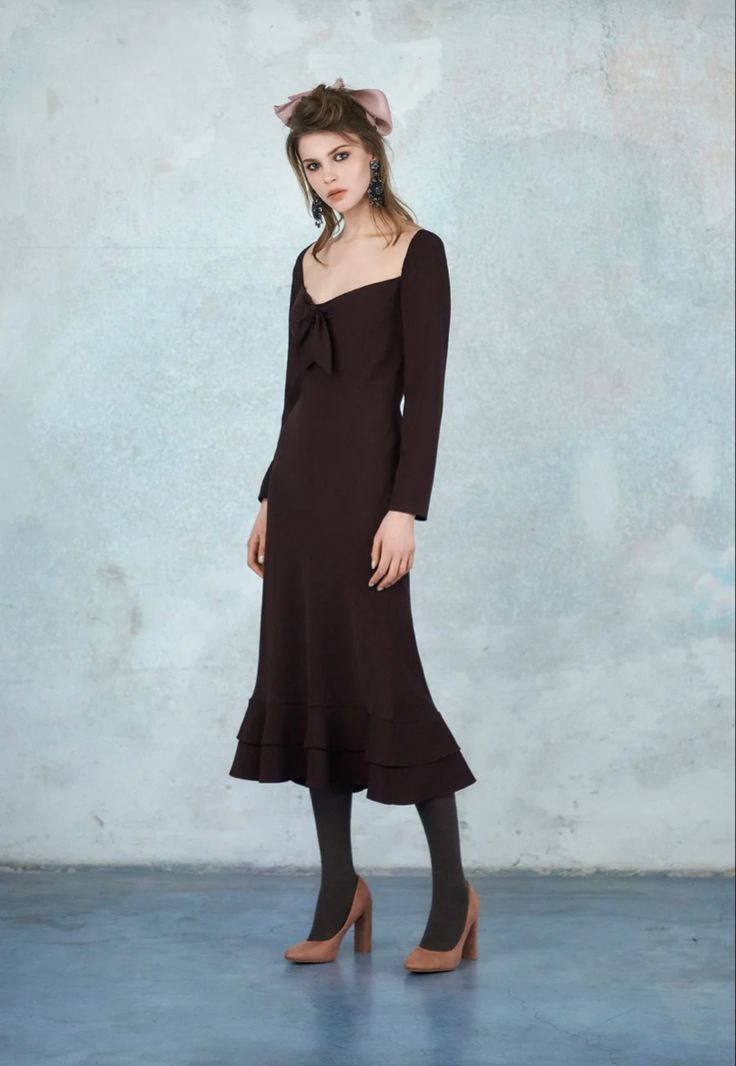 a fulllength studio shot of a young woman wearing a dark brown midi dress. the dress is a longsleeve, squareneck style with a tie at the neckline, and has a ruffled hemline.  she is also wearing brown heeled pumps, a light pink hair bow, and earrings. the woman is standing against a textured, light blue wall.