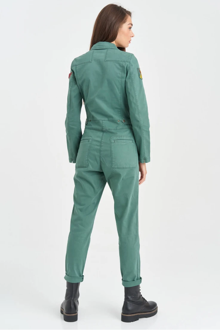 a woman in a green jumpsuit and black boots poses against a white backdrop. she is looking off to the side and her hair is pulled back from her face. the jumpsuit is casual and flattering. the woman looks stylish and confident.