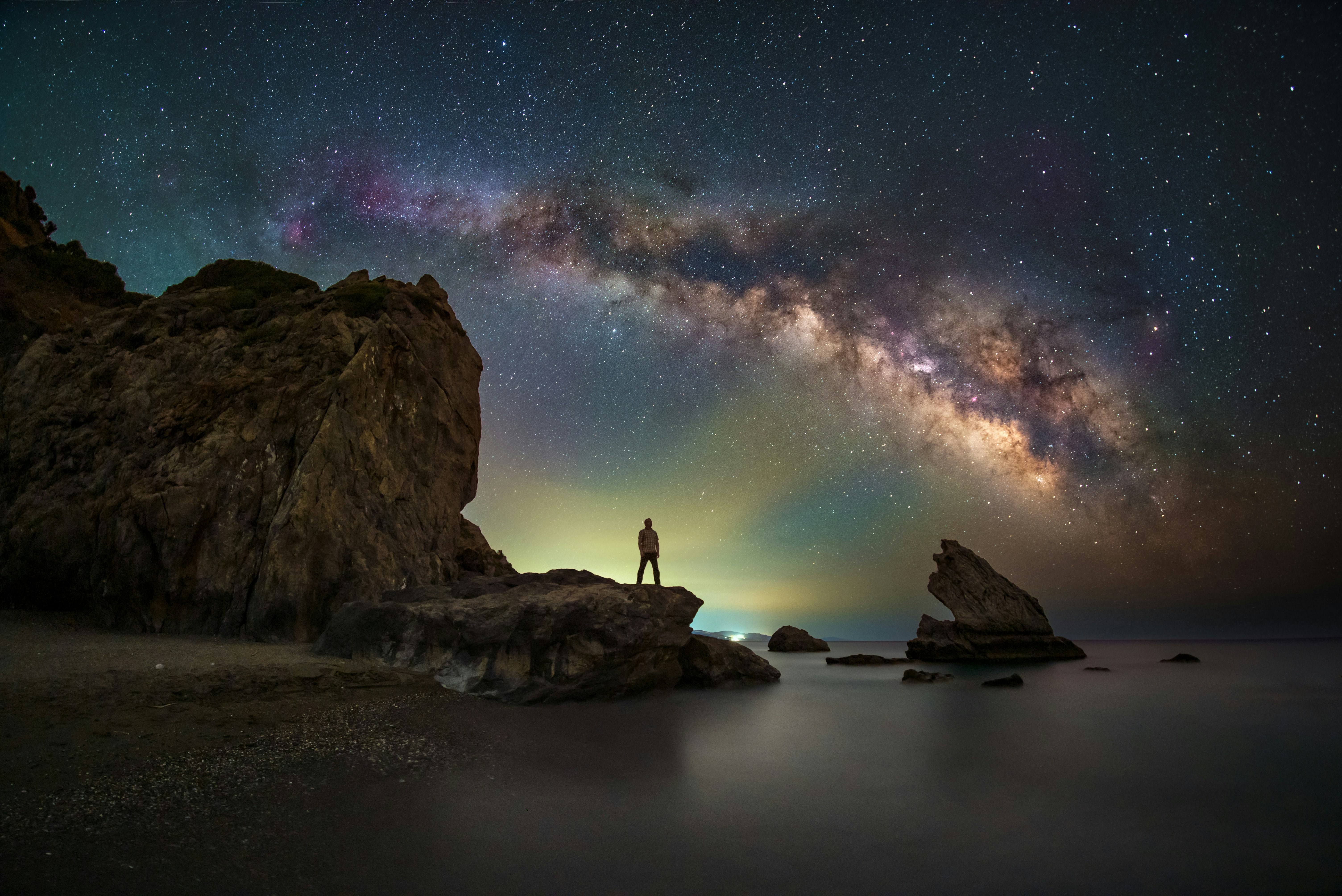 a breathtaking wideangle astrophotography image captures the milky way galaxy arching across a night sky. a silhouetted man stands on a rocky outcrop at the edge of a calm ocean, gazing up at the spectacle above.  the long exposure smooths the water's surface, reflecting the faint glow of distant lights and the starry sky.  two other large rock formations rise from the sea, creating a dramatic foreground against the cosmic backdrop. the scene evokes feelings of awe, wonder, and the insignificance of humanity against the vastness of the universe.