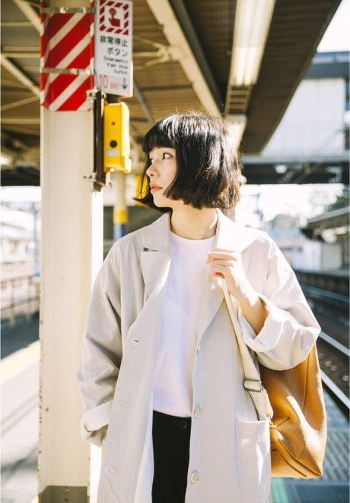a young woman with a chic bob haircut stands on a train platform bathed in warm sunlight. she's stylishly dressed in a light grey trench coat layered over a white tshirt and dark pants. a brown leather tote bag hangs from her shoulder. she's turned to the side, seemingly looking down the tracks, possibly waiting for the train. the red and white striped signage and glimpse of the train tracks hint at a japanese train station setting.