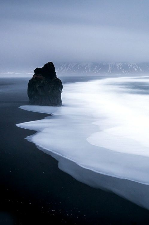 a long exposure shot capturing a basalt rock formation on the black sand beach of reynisfjara in vik, iceland. the ocean waves swirl around the rock, creating a smooth, ethereal effect. a misty mountain range is visible in the background under an overcast sky. the overall mood is dramatic and moody.