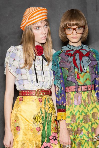 two young women models are showcased in a fashion show, likely for gucci, given the style of clothing.  the model on the left sports an orange striped beret, a light purple and white checkered blouse, a red studded gucci belt, and a vibrant yellow skirt with floral embroidery. her hair is long and styled with bangs. the model on the right has a shorter bob haircut with bangs, wears glasses, and a vibrant multicolored blouse and skirt featuring a mix of patterns and colors.  both outfits feature bright, bold colours and patterns, indicative of a spring/summer collection. the background is a dark, muted grey, providing contrast to the bright clothing.