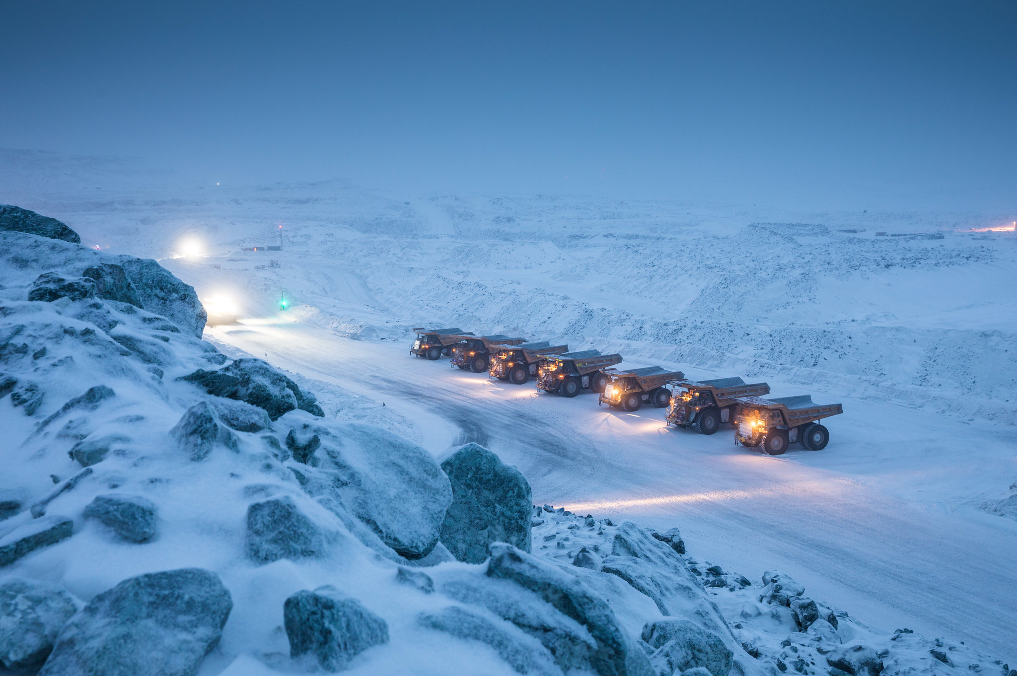 a line of large yellow dump trucks drive down a snowcovered haul road in a remote mine site. the trucks are driving in a single file line, and they are all carrying large loads of ore. the landscape is barren and rocky, and the snow is deep and powdery. the sky is blue, and the sun is setting.