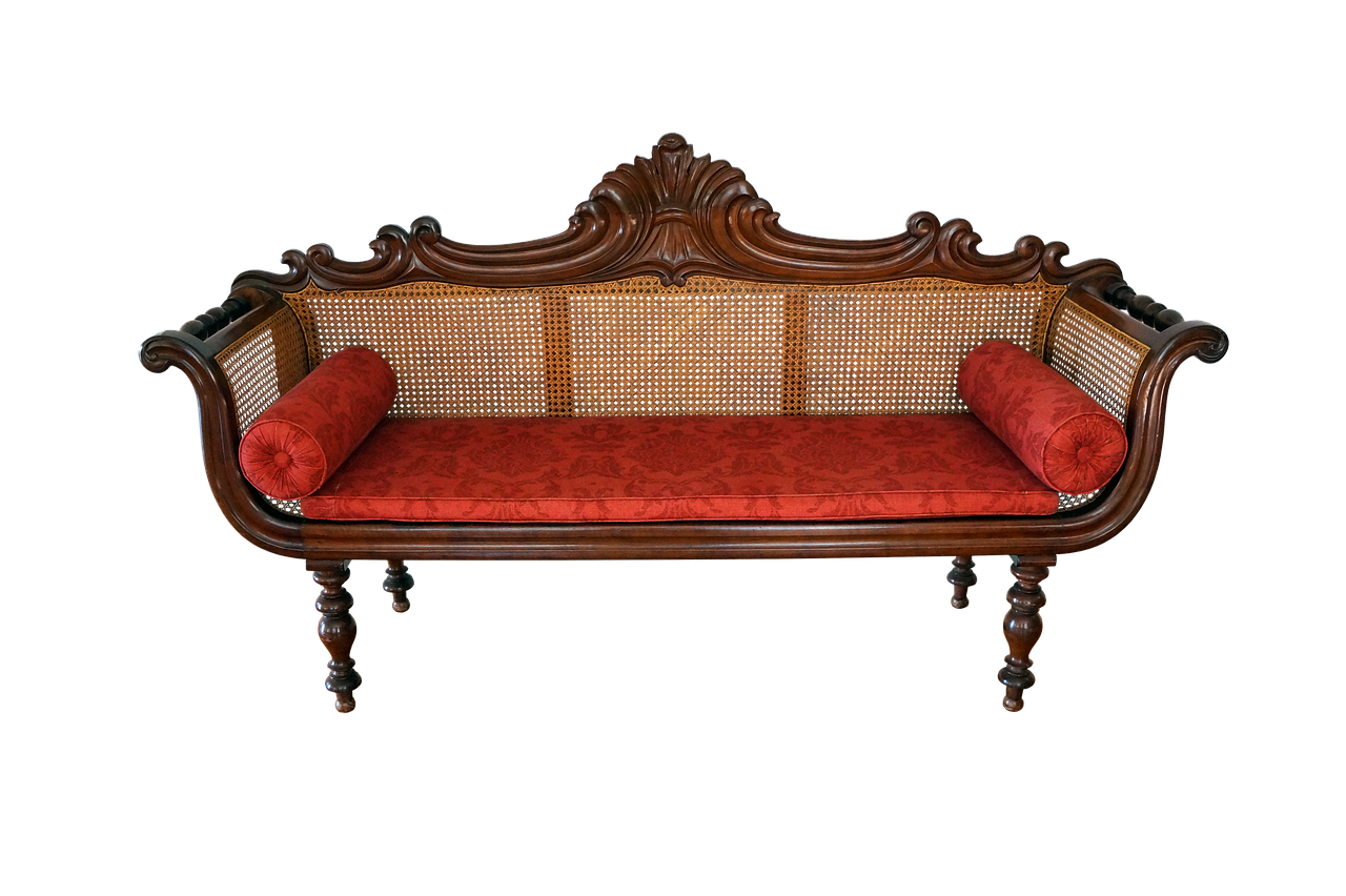 an antique wooden sofa with a cane back and ornate carving details, featuring red cushions and bolster pillows, set against a black background.