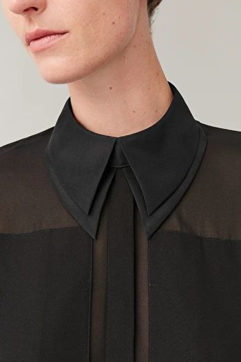 loseup shot of a woman's black sheer shirt with a unique, sharplypointed collar. the shirt is semitransparent, revealing a hint of the model's skin underneath. the collar is a prominent feature, crafted from a heavier black fabric than the shirt itself. the image focuses on the details of the shirt and collar, showcasing the texture and design. the overall aesthetic is elegant and sophisticated. the background is a neutral, light gray.  the model's face is partially visible, but the focus is primarily on the clothing.