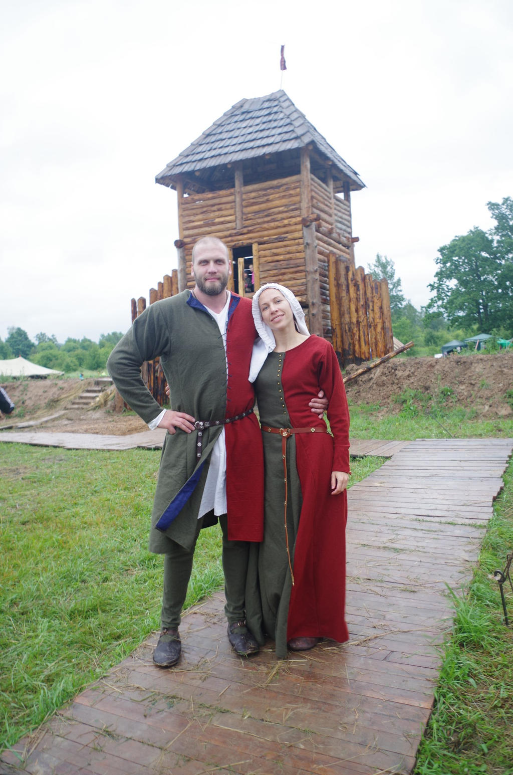 a man and woman, both dressed in medieval clothing, pose on a wooden pathway in front of a wooden tower. the man is wearing a long green tunic with blue trim, a white undershirt, and brown boots. he has a belt around his waist and one hand on his hip. the woman is wearing a long red and green dress with a white head covering. the wooden tower is made of logs and has a small roof. the background is a grassy field with some trees.
