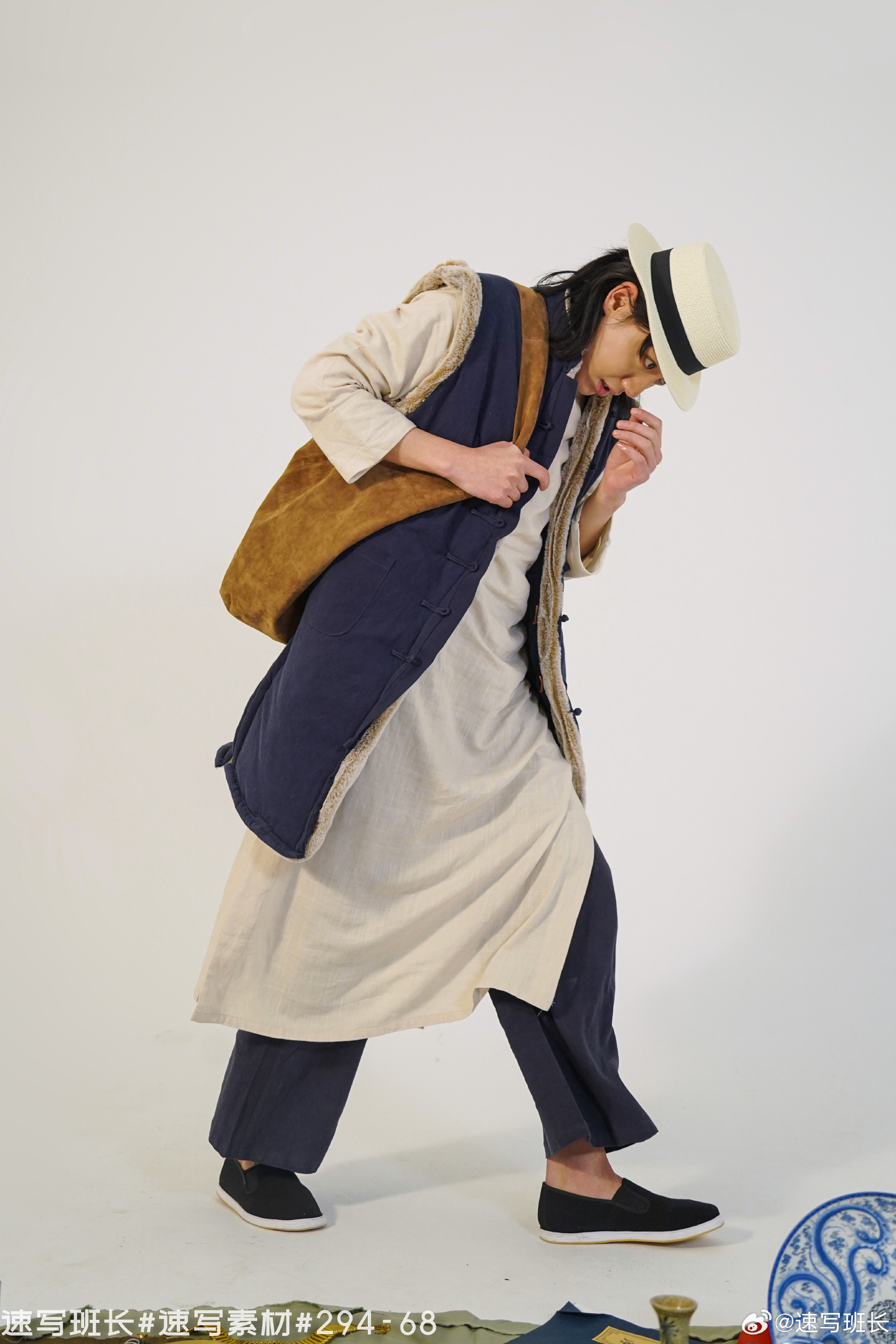 a man wearing a long, lightcolored robe, dark blue pants, a blue vest with brown fur lining, a brown shoulder bag, and a straw hat is standing in front of a white background. he is looking down and adjusting his collar.  he is wearing darkcolored slipon shoes. there are hints of colorful fabrics and metallic decorative items on the floor around his feet.