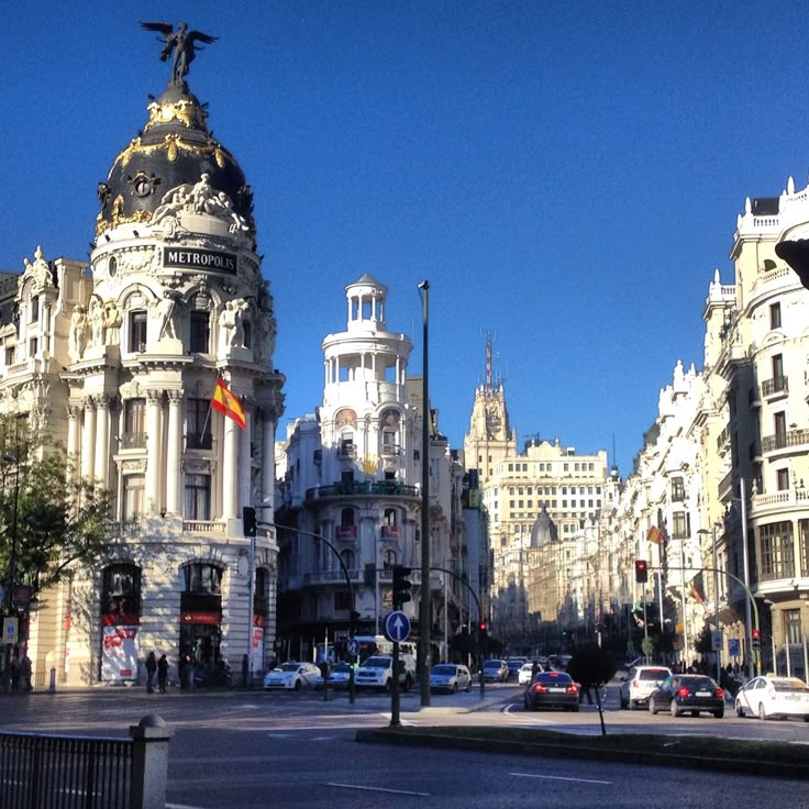 the image shows a vibrant street scene in madrid, spain, specifically on gran vía. the iconic metropolis building dominates the left side of the frame, its ornate architecture standing out against the clear blue sky. the street is lined with other elegant buildings, typical of madrid's architecture.  cars are visible on the street, and a few people are walking along the sidewalks. the overall impression is one of a bustling yet beautiful european city on a bright, sunny day. the spanish flag is visible fluttering from a pole on the metropolis building.