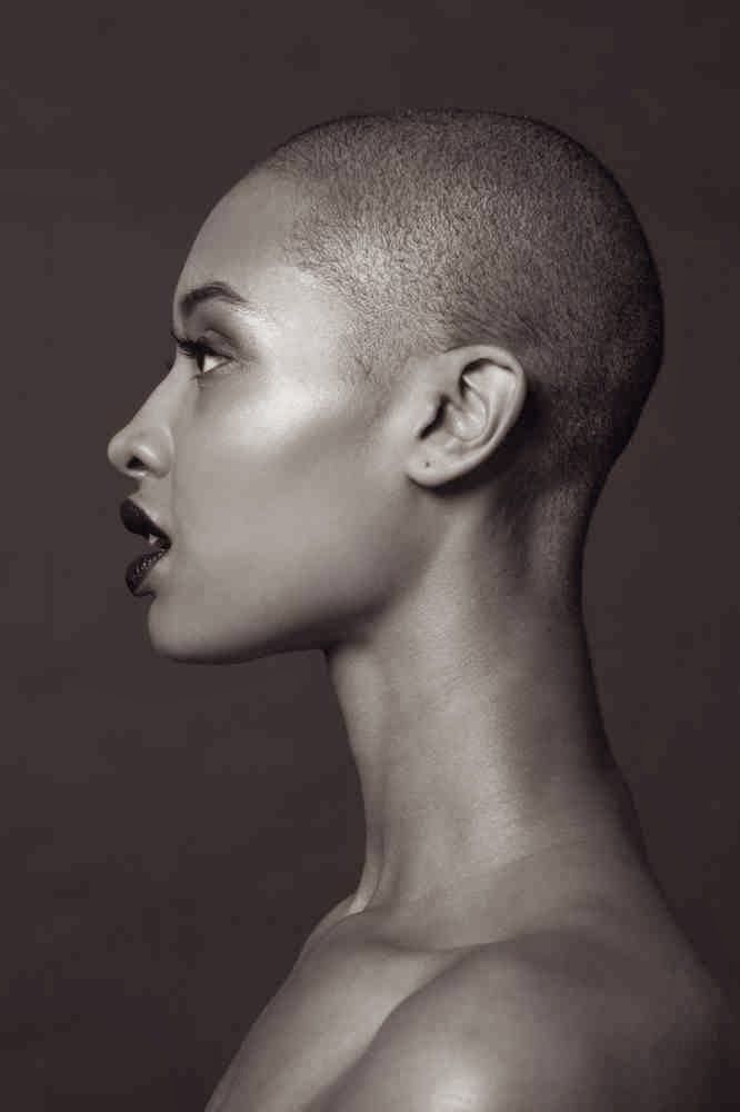 a black and white portrait shows a woman in profile with a shaved head, facing left. she has dark lipstick and a serious expression. the image highlights her neck and bare shoulders, with a focus on skin texture and the monochrome aesthetic.