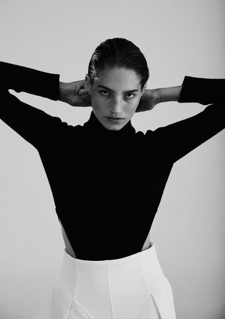 a young woman in a black turtleneck top and white high waisted pants is standing in a studio with a white background. she is looking directly at the camera with a serious expression on her face.  her short hair is slicked back, and her hands are placed on her shoulders. the lighting is soft, and the image is in black and white.