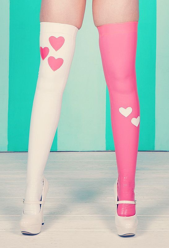 a pair of legs wearing mismatched stockings and shoes are shown. the stocking on the left leg is white with three pink hearts, and the shoe is white. the stocking on the right leg is pink with two white hearts, and the shoe is white. the shoes are mary janes with a platform heel. the background is a light teal with vertical dark teal stripes.