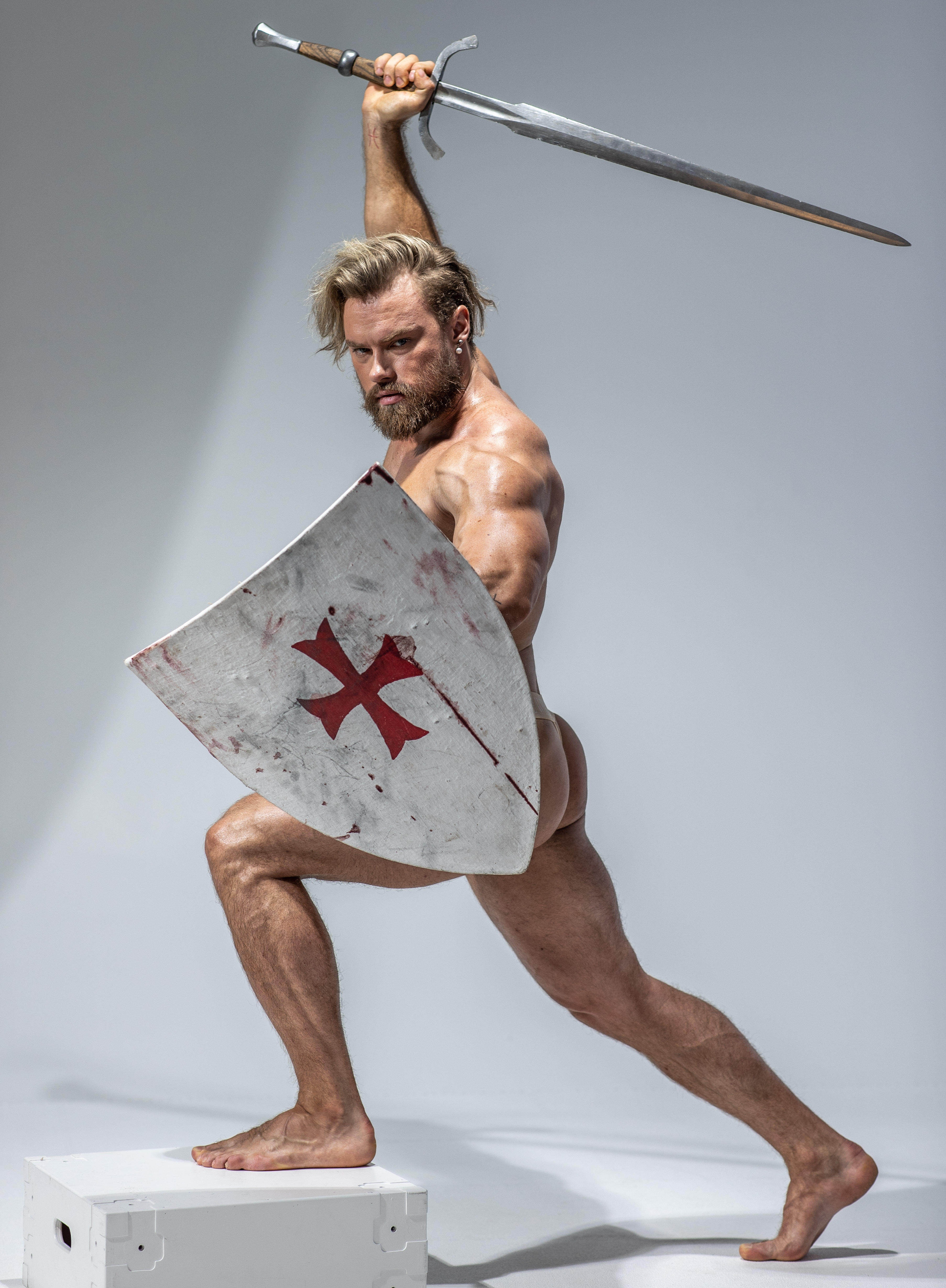 a muscular man, nude and holding a sword and shield, poses dynamically against a light grey studio backdrop. he has a beard and longish blond hair and stands with one foot on a white box, suggesting motion. the shield bears a red cross, hinting at a crusader or templar knight theme.  the scene evokes historical warfare and highlights themes of strength and power.