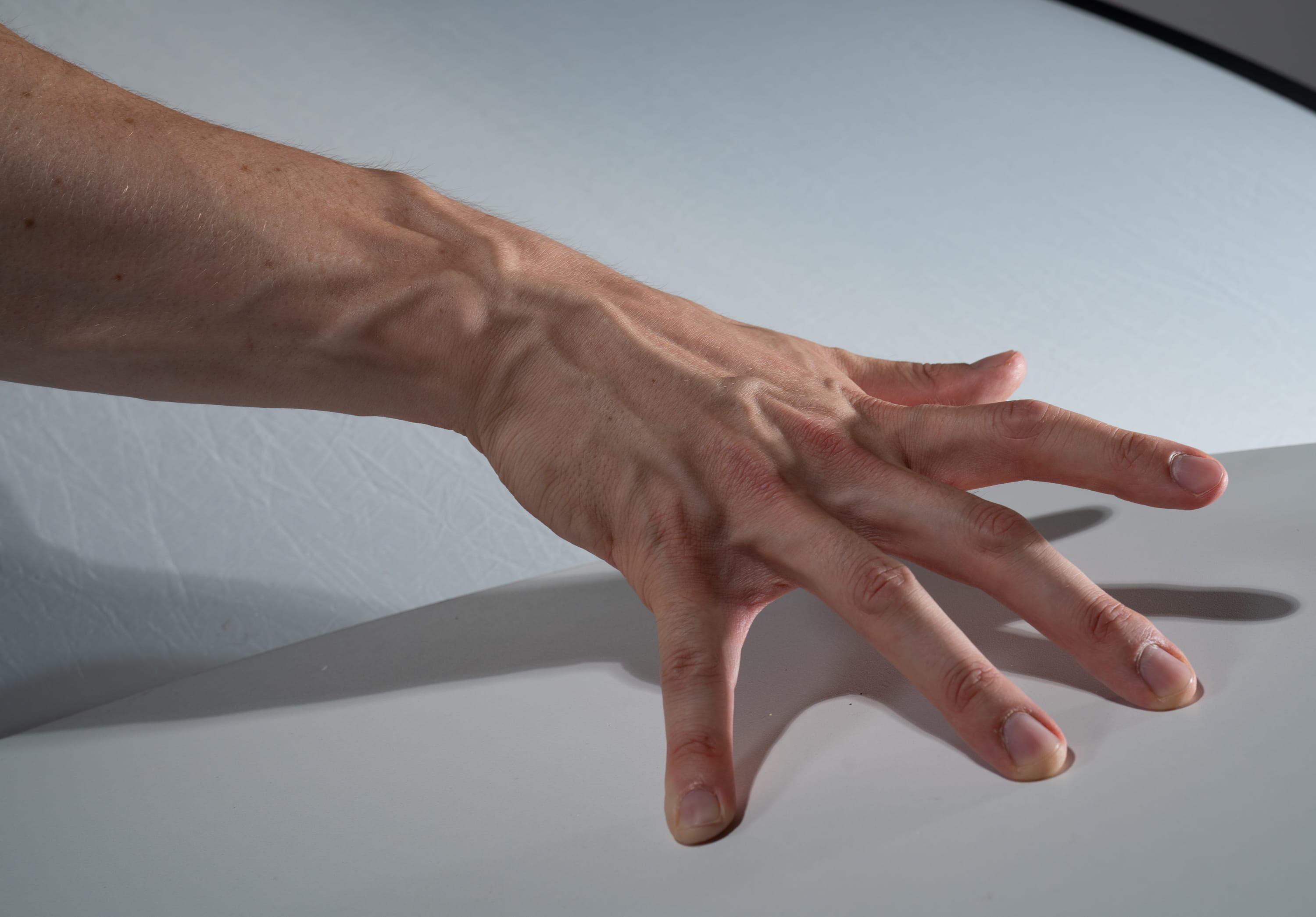 a closeup shot of a hand with prominent veins pressed against a white surface. the hand is positioned with fingers spread out, showcasing the details of its structure and the network of veins. the lighting is bright, likely studio lighting, highlighting the textures and shadows on the hand.