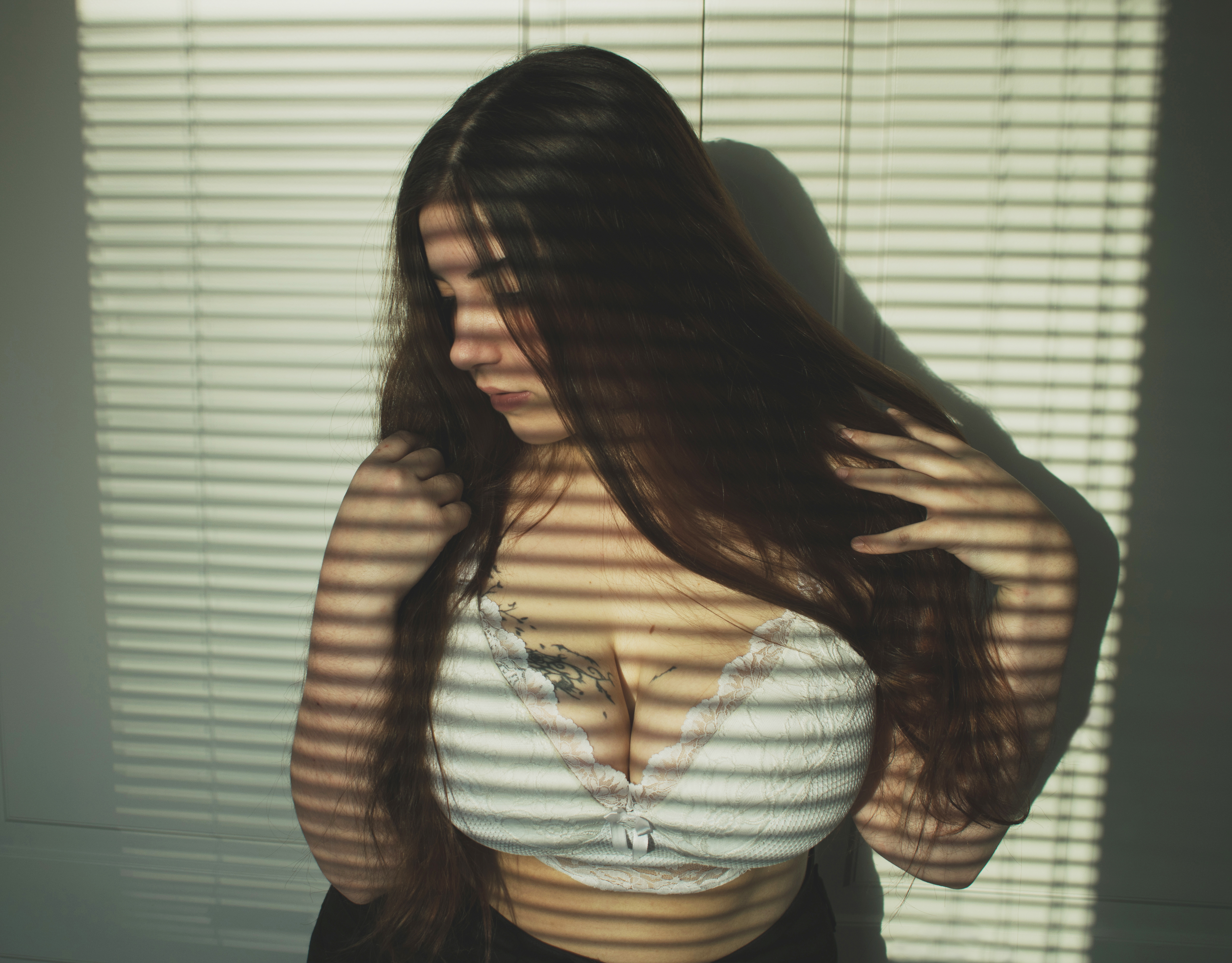 a woman with long brown hair stands in front of blinds that cast shadows across her face and body. she is wearing a white bra and a dark bottom. she is looking down, with one hand resting on her chest and the other touching her hair. a tattoo is visible on her chest.