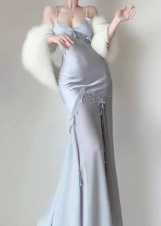 a woman with long blonde hair is wearing a long blue satin dress with a fur wrap, posing with her arm raised and looking away from the camera. she is wearing pearl earrings and a thin necklace. she is standing in a welllit room with a white background.