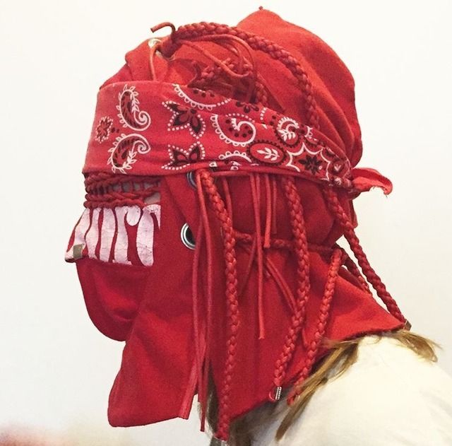 a closeup shot of a person wearing a red bandanastyle mask with a large red hood. the mask is adorned with braids and strings, and the hood has a zipper down the front. the background is white, and the person's hair can be seen peeking out from under the hood.