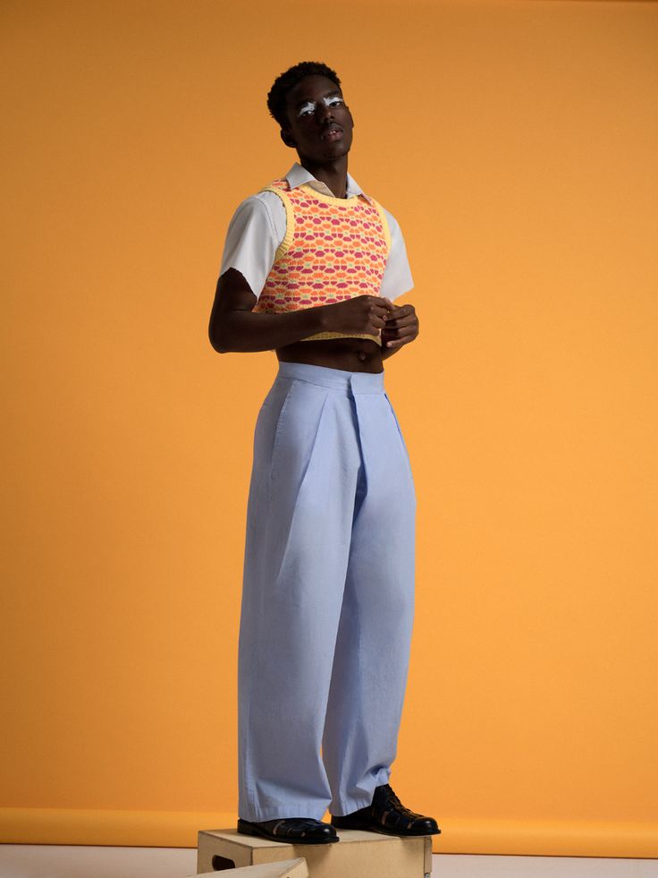 young male model stands on a small box against an orange backdrop. he is wearing a shortsleeved white shirt layered under a cropped, brightly colored, patterned knit sweater vest. his trousers are widelegged and light blue.  his shoes are darkcolored and he is striking a pose.  the image is fashion photography, with a focus on the clothing.