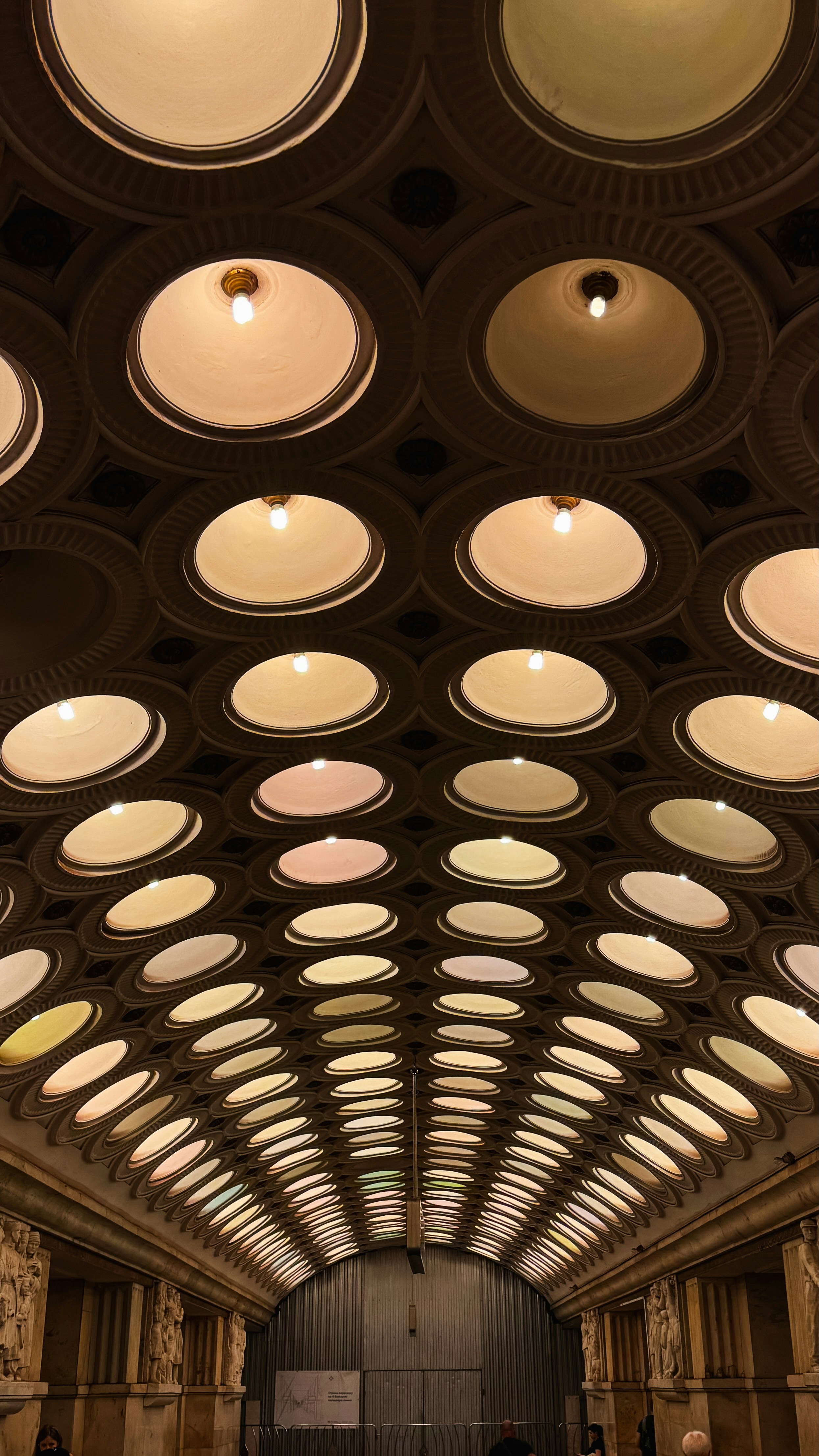 the image captures a lowangle perspective of an ornate hallway. the focal point is the intricately designed ceiling adorned with rows of circular, recessed lights, creating a captivating symmetrical pattern. the warm, artificial light from these fixtures illuminates the space, highlighting the architectural details and casting soft shadows. the hallway narrows towards the end, leading to an arched doorway with a metal gate. two individuals can be seen at the far end of the hall. the overall ambiance is one of grandeur and elegance, characteristic of a subway station with a focus on architectural splendor.
