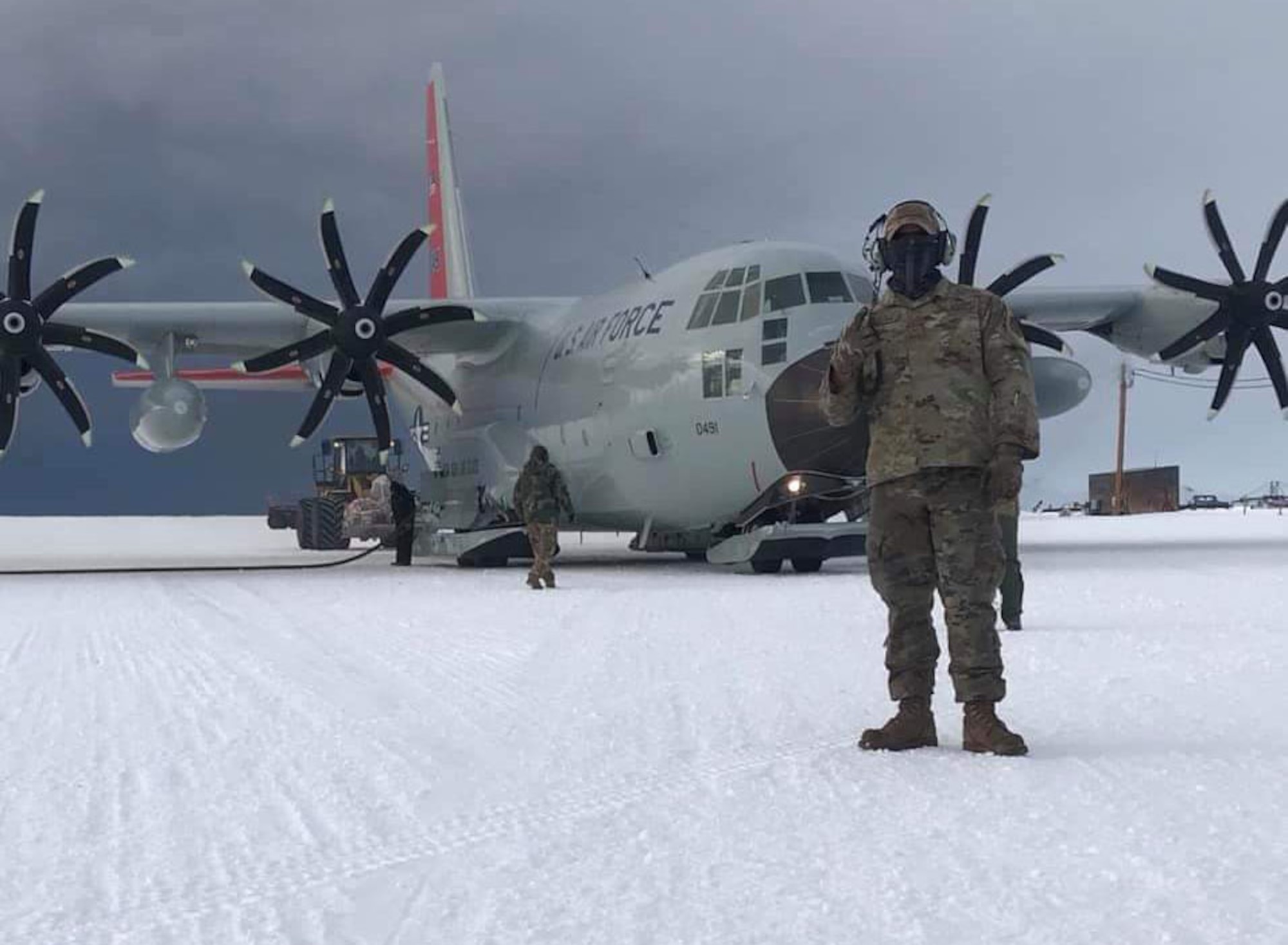 a us air force c130 cargo plane sits on a snowy runway with a soldier standing in front of it. the plane is being loaded with fuel and supplies.  a truck can be seen in the background.  there is a small group of people in the foreground, presumably ground crew. the plane is ready for its next mission in the arctic.