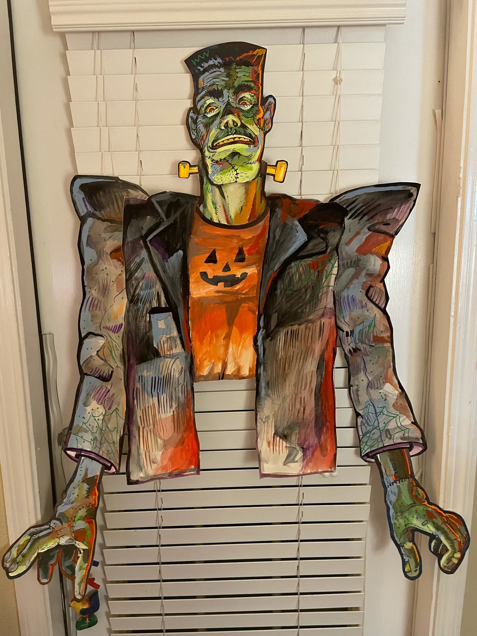 the image shows a vibrant, handpainted cardboard cutout of frankenstein's monster hanging in a window.  the monster is depicted in a slightly cartoonish, pop art style, wearing an orange shirt with a jacko'lantern design and a multicolored jacket.  the artwork is quite detailed, with visible brushstrokes and a blend of colors. the cutout is suspended by strings in front of white blinds, suggesting it's a halloween decoration. the style is expressive and playful rather than strictly realistic.