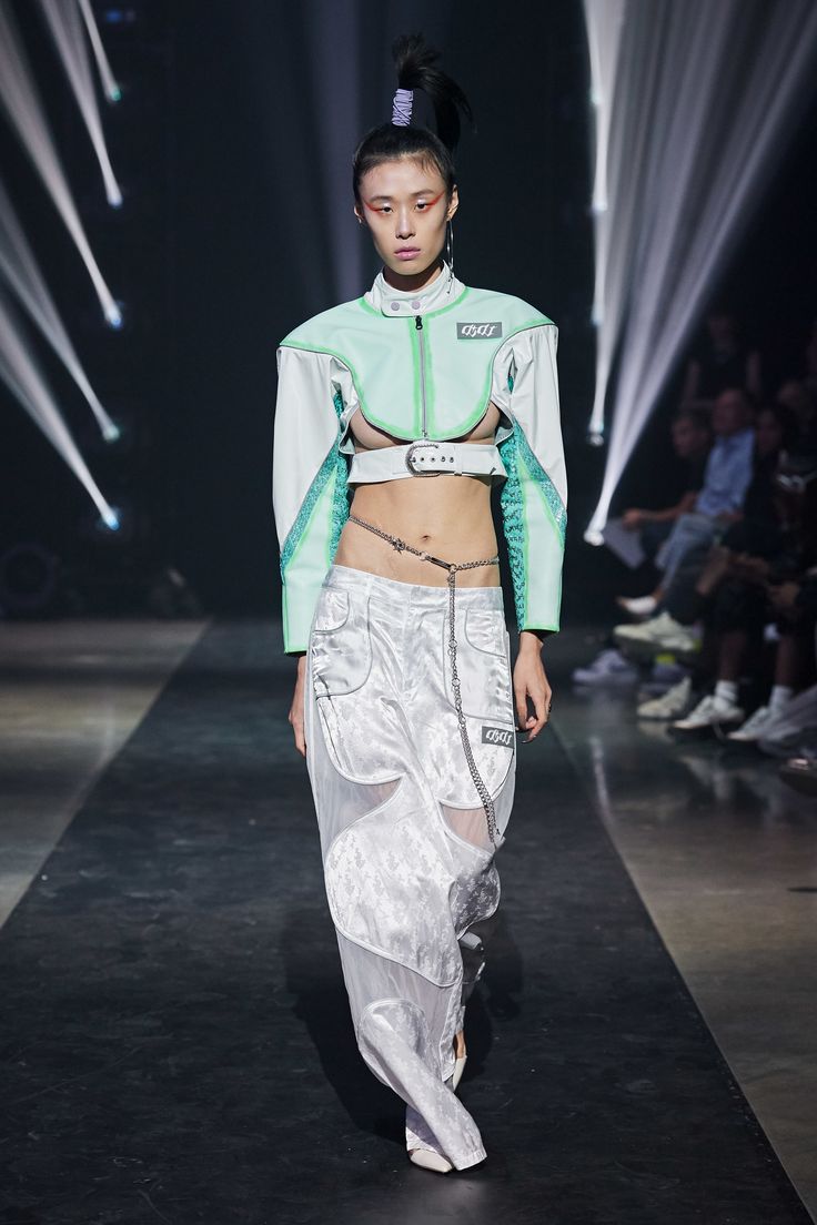 a female model walks the runway in a unique, futuristic outfit. the model has a long black ponytail and is wearing a green, white, and black outfit. she is wearing a cropped, green and white jacket that exposes her midriff. the jacket is cinched at the waist with a black belt.  she is also wearing a pair of white, baggy, pants that have a black chain attached to them. the model is also wearing a necklace that wraps around her waist. the model is posing for the camera, and the background is blurred.