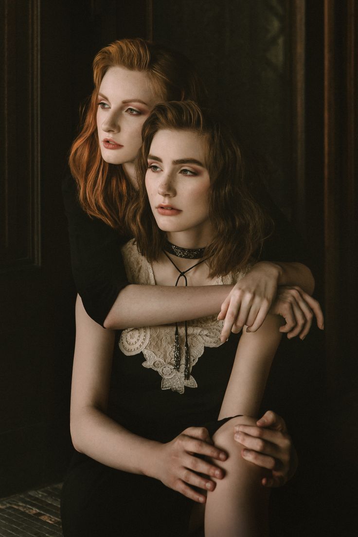 two women are sitting close together, their arms wrapped around each other. they are wearing black dresses, the one in the front has a lace