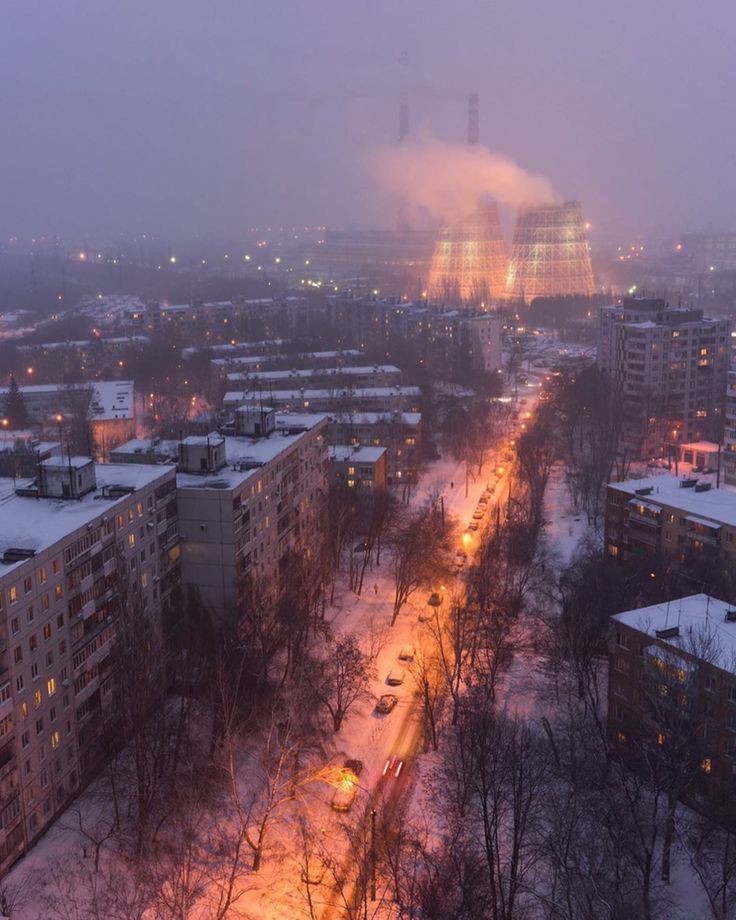 an aerial shot of a city at twilight in winter.  the scene is softly lit by streetlights and the glow of buildings. snow covers the ground and trees. in the background, large industrial cooling towers emit smoke or steam into the dusky sky. cars are visible driving along a street that winds through the residential area. the overall mood is atmospheric and slightly melancholic.