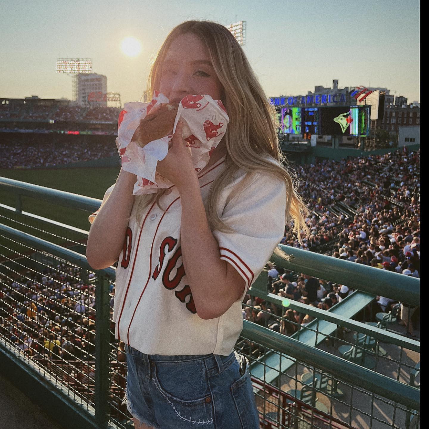 the image shows a young blonde woman wearing a creamcolored boston red sox baseball jersey and denim shorts. she is standing at the railing of fenway park, a wellknown baseball stadium, during a game. a large crowd of spectators fills the stands behind her.  the sun is setting, casting a warm glow.  the woman is happily eating what appears to be a hot dog wrapped in paper. the overall mood is relaxed and joyful, capturing a quintessential summer day at a baseball game.