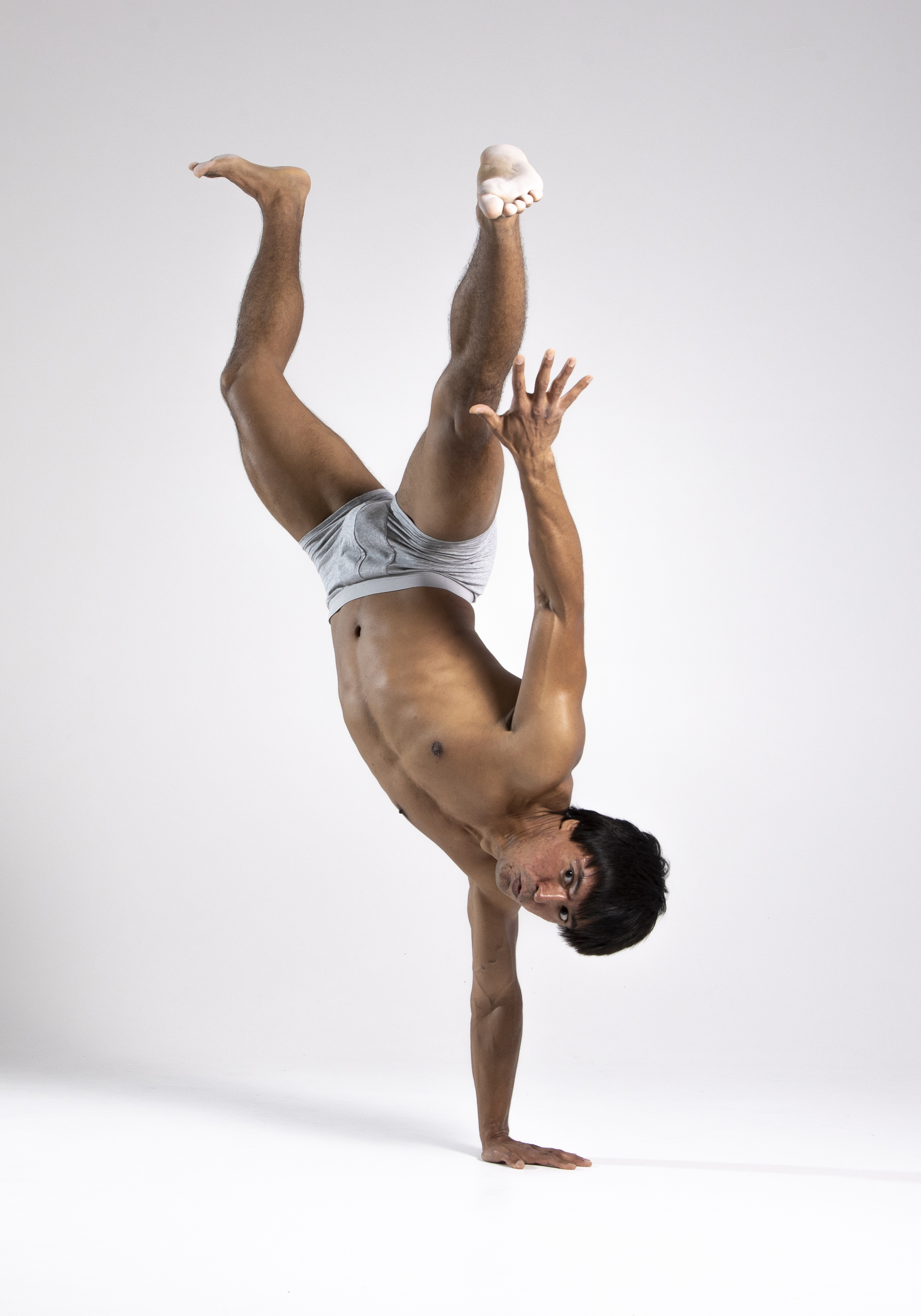a man is performing a handstand in a studio. he is shirtless and wearing underwear. one leg is straight up in the air and the other is bent at the knee. he is looking towards the camera with one hand on the floor and the other up in the air. he is barefoot. the background is a plain white studio background.
