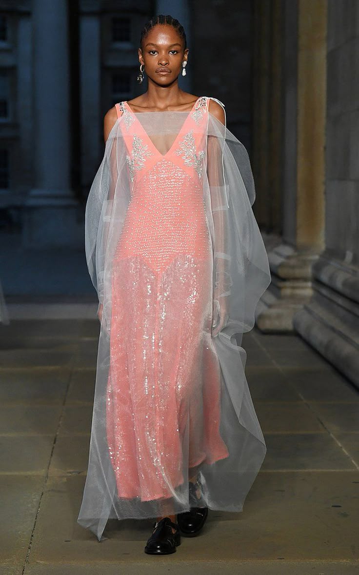 a black woman stands on a runway, wearing a light peach sequin dress with a deep vneckline and a floorlength skirt, covered with a sheer tulle veil. she also has on black shoes and earrings. the background features columns and an indoor setting.