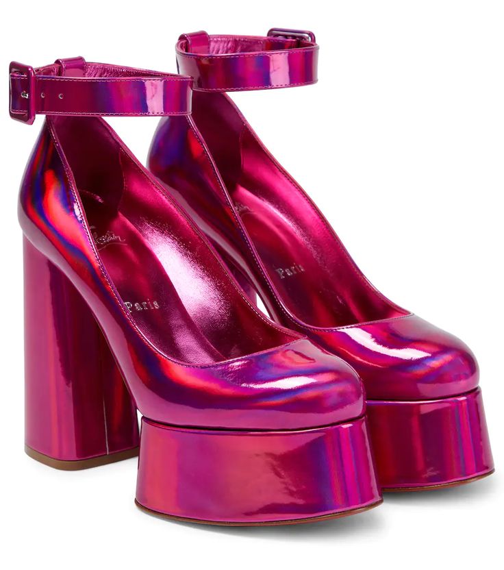 a pair of pink metallic platform high heels with ankle straps are displayed on a white surface. the shoes feature a round toe, thick platform sole, and chunky block heel. the ankle straps have a buckle closure. the inside of the shoe is pink and marked with "paris." the shoes have a glossy, reflective finish.