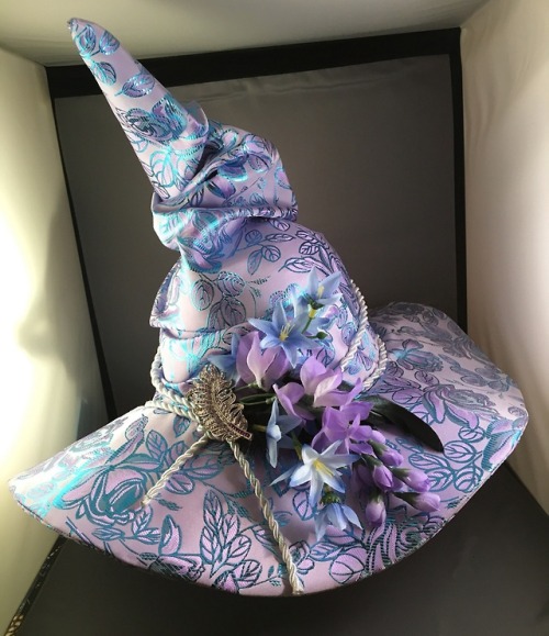 a purple and blue floral witch hat with a tall, pointed crown. the hat is decorated with a silver leaf brooch and a spray of purple and white flowers. the hat is made of a brocade fabric and has a wide brim. it is displayed on a white surface in a welllit room.