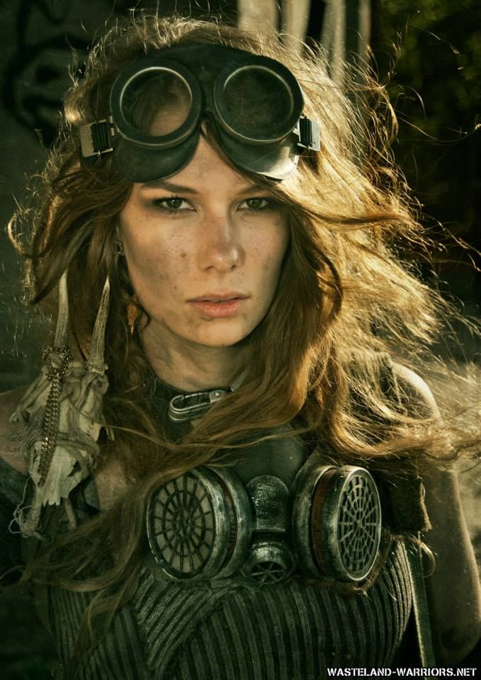 a closeup portrait of a woman with long, messy blonde hair wearing futuristic goggles and a gas mask. she is dressed in a dirty, striped shirt and leather collar with a worn necklace and earrings. the background is blurred and out of focus. the woman is looking directly at the camera with a serious expression.