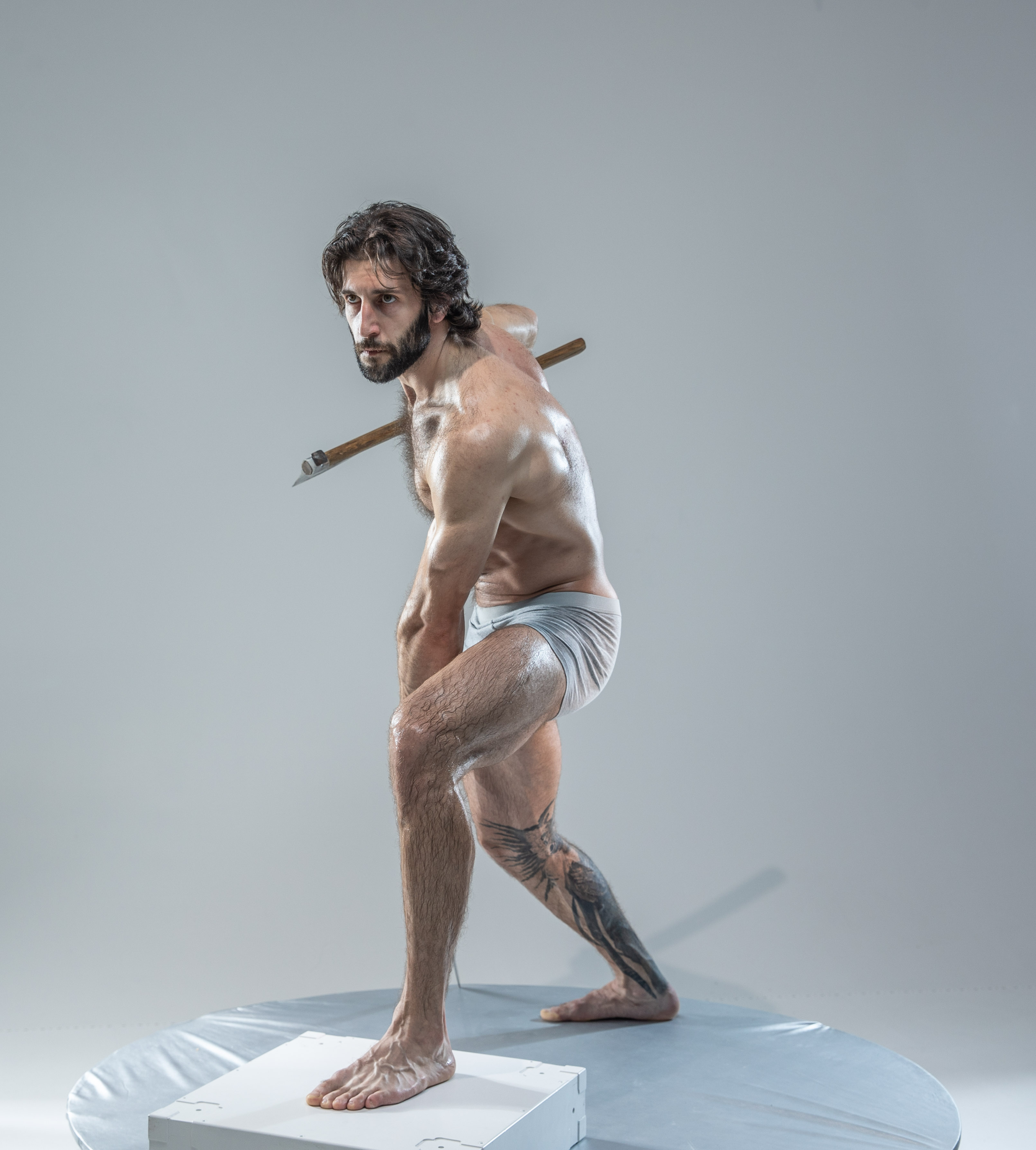 a muscular man with a beard and tattoos stands on a white platform in a studio setting. he is wearing only white underwear and holds a wooden axe in a defensive posture. the image evokes strength, power, and a warriorlike aesthetic. his intense gaze and the dynamic pose further accentuate the feeling of action. the lighting is soft and diffused, highlighting the man's physique. the overall color palette is predominantly gray, with the man's tanned skin providing a contrast.