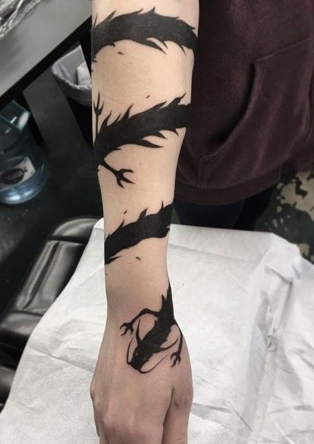a closeup image of a person's arm with a black dragon tattoo. the tattoo is done in a linework style and is very detailed. the tattoo starts on the forearm and goes up to the wrist. the person's hand is visible and the dragon appears to be wrapping around their hand. the tattoo is done in black ink and is very intricate. the background is blurry, but it looks like the person is sitting on a couch.