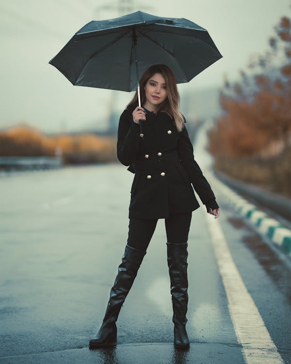 a fulllength shot of a young woman standing on a wet road, holding a dark umbrella. she's dressed in a stylish black doublebreasted coat, black leggings, and thighhigh black boots. the background is blurred, showing autumnal foliage and a grey sky, giving the scene a moody, atmospheric feel. the woman's pose is relaxed yet elegant as she looks directly at the camera. the overall image is wellcomposed, with a focus on the woman and her fashionable attire.
