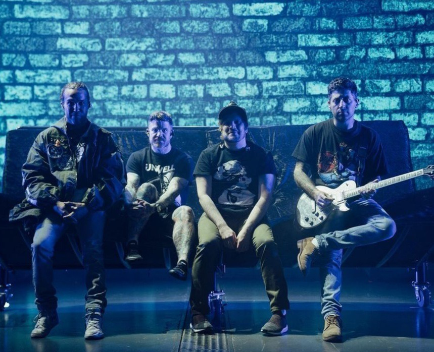 a rock band of four men are sitting on a stage in front of a blue brick wall. the band members are playing their instruments. the band members are all dressed in casual clothes, and they all have microphones in front of them. they are all looking at the camera.  one man is playing a guitar while sitting, another is sitting on the edge of the stage with a microphone and no instrument, and two more men are sitting on a bench, with one playing a guitar and the other holding a microphone.