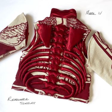 a closeup shot of a unique handmade garment with a red skeleton design on the front. it's a red and white fabric with removable sleeves and a bold, oversized design. the fabric has a textured appearance, and the piece is clearly made with attention to detail and craftsmanship. the word "removable sleeves" is handwritten in black ink on the bottom left, and the word "marie iv" is handwritten in black ink on the top right. this piece appears to be a oneofakind garment that is sure to turn heads.