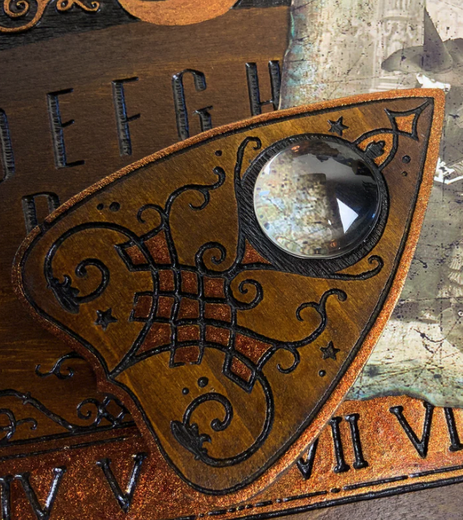 a closeup shot of a wooden ouija board planchette. it is carved with intricate details, featuring a black outline and a goldlike paint finish. the planchette is set against the background of a wooden ouija board featuring roman numerals and the letters defg.