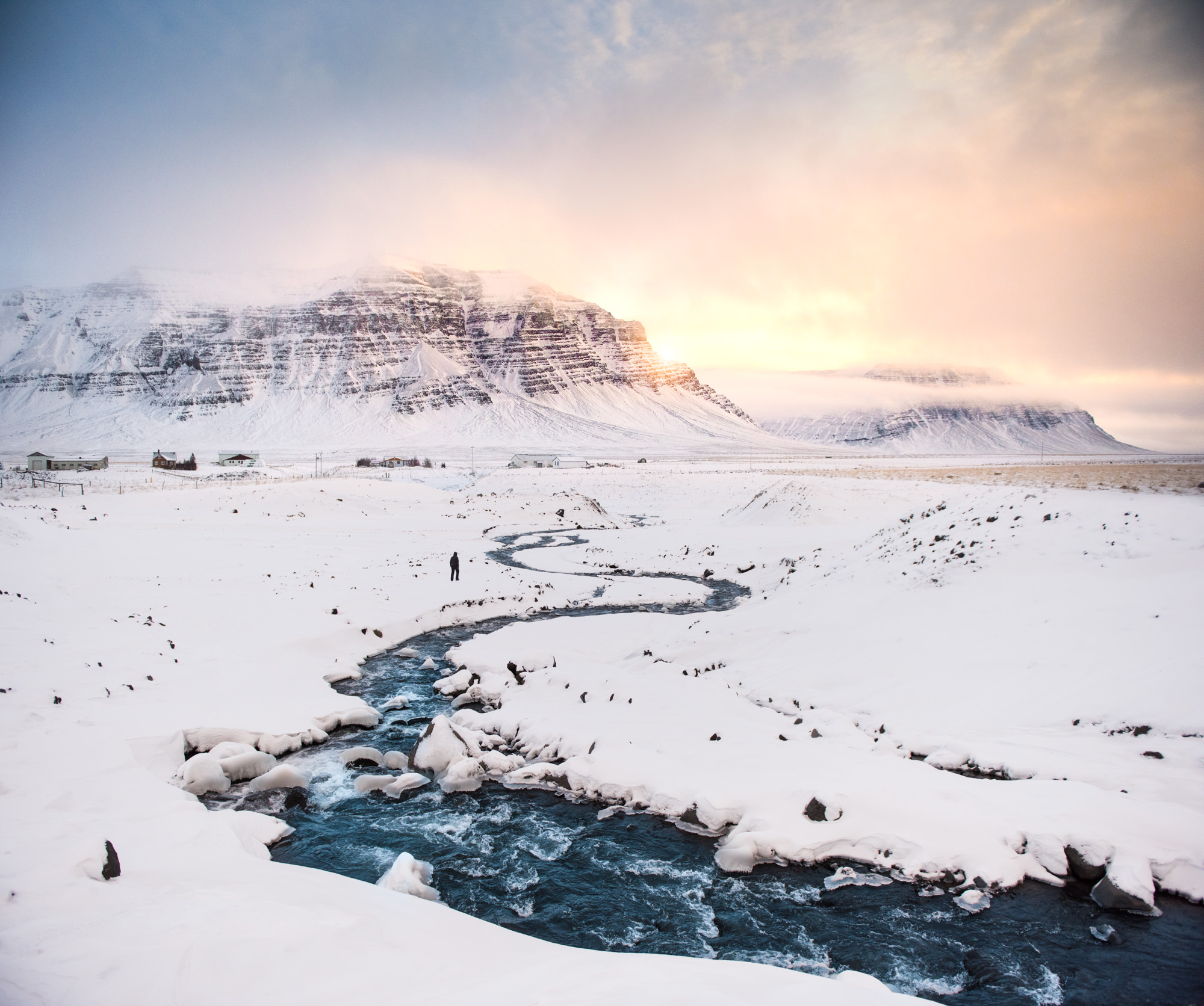 a snow covered mountain landscape with a winding river flowing through it. the sun is setting over the mountains and casting a warm glow over the scene.  the snow is pristine and the river is clear and blue.  the image is serene and peaceful.
