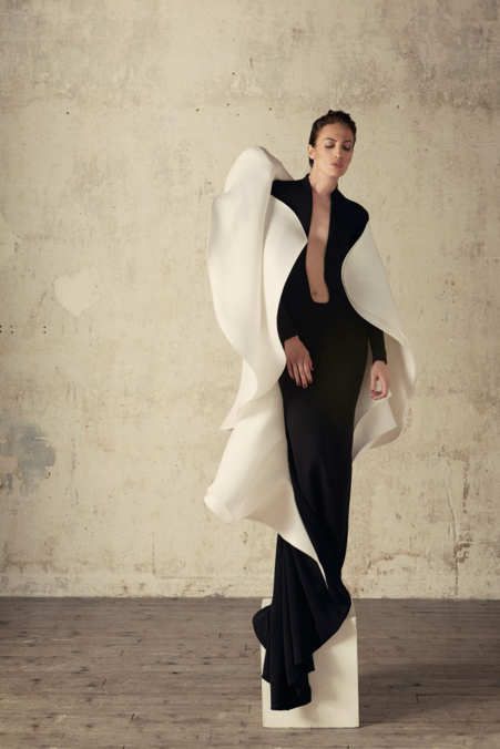 a full shot captures a woman in a striking black dress with a plunging neckline, complemented by a large, sculpted white cape. she stands on a white pedestal in a studio setting against a textured beige backdrop. the image is a fashion portrait.
