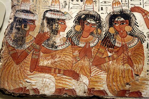 the image shows a painting of four ancient egyptian women, likely musicians or performers. they are depicted sitting sidebyside, wearing long dresses and elaborate headdresses. the woman on the far right is playing a flutelike instrument. the painting is done in warm, earthy tones and features intricate details.